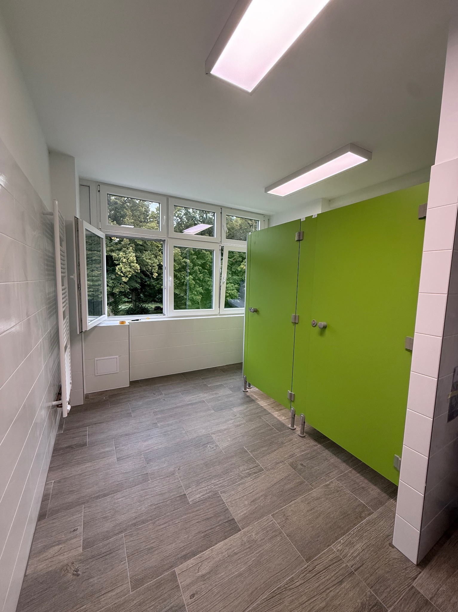 Nowoczesna toaleta publiczna z zielonymi kabinami, szarą podłogą imitującą drewno i dużym oknem z widokiem na zieleń. Minimalistyczny design, jasne oświetlenie.