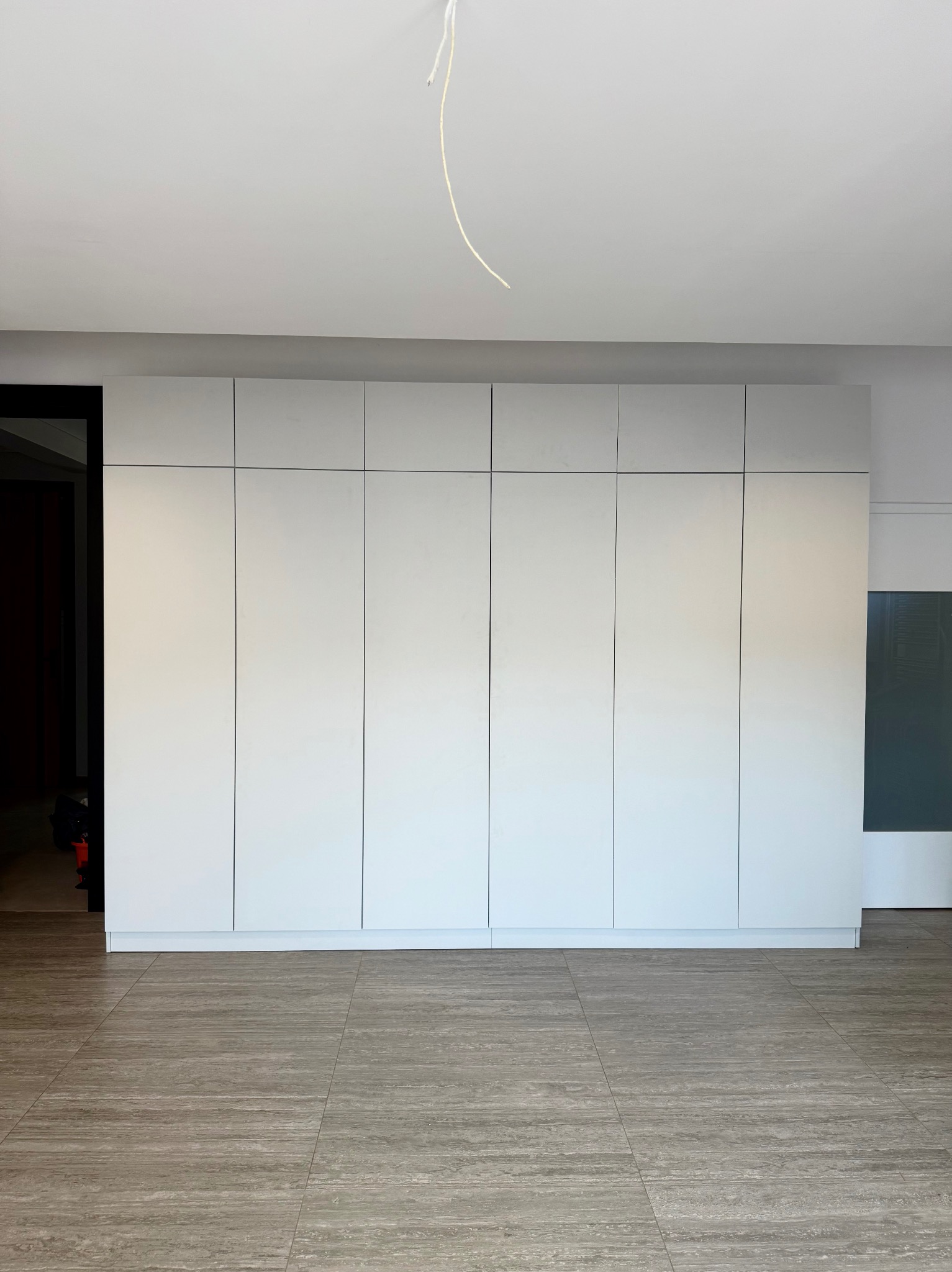 Biała, minimalistyczna szafa wnękowa z siedmioma drzwiami, na tle szarej podłogi imitującej drewno. Widoczny fragment instalacji elektrycznej na suficie.