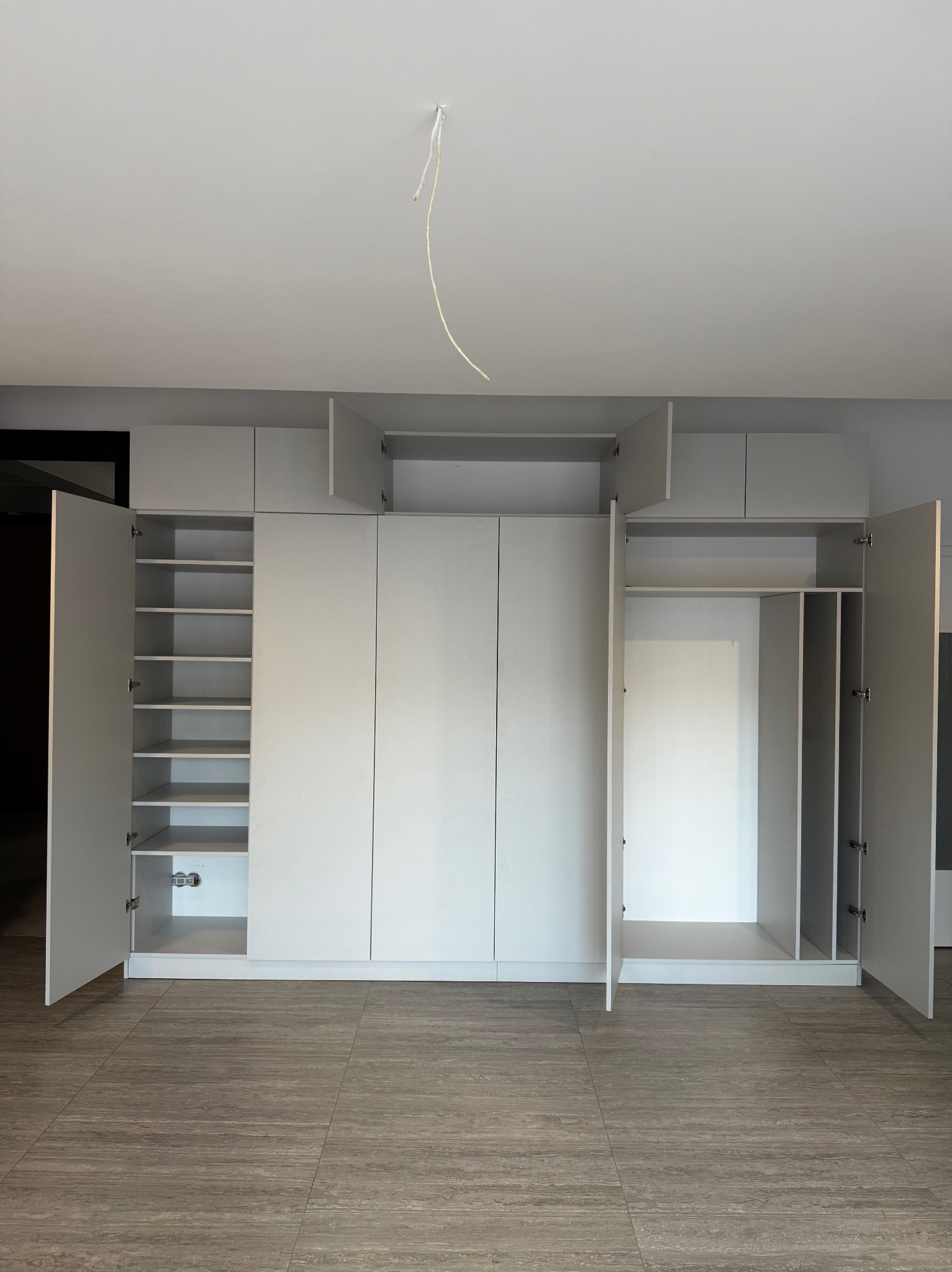 Jasnoszara szafa na wymiar z otwartymi drzwiami, ukazująca półki i przestrzeń do wieszania ubrań. Widoczne szare płytki podłogowe i wystający kabel z sufitu.