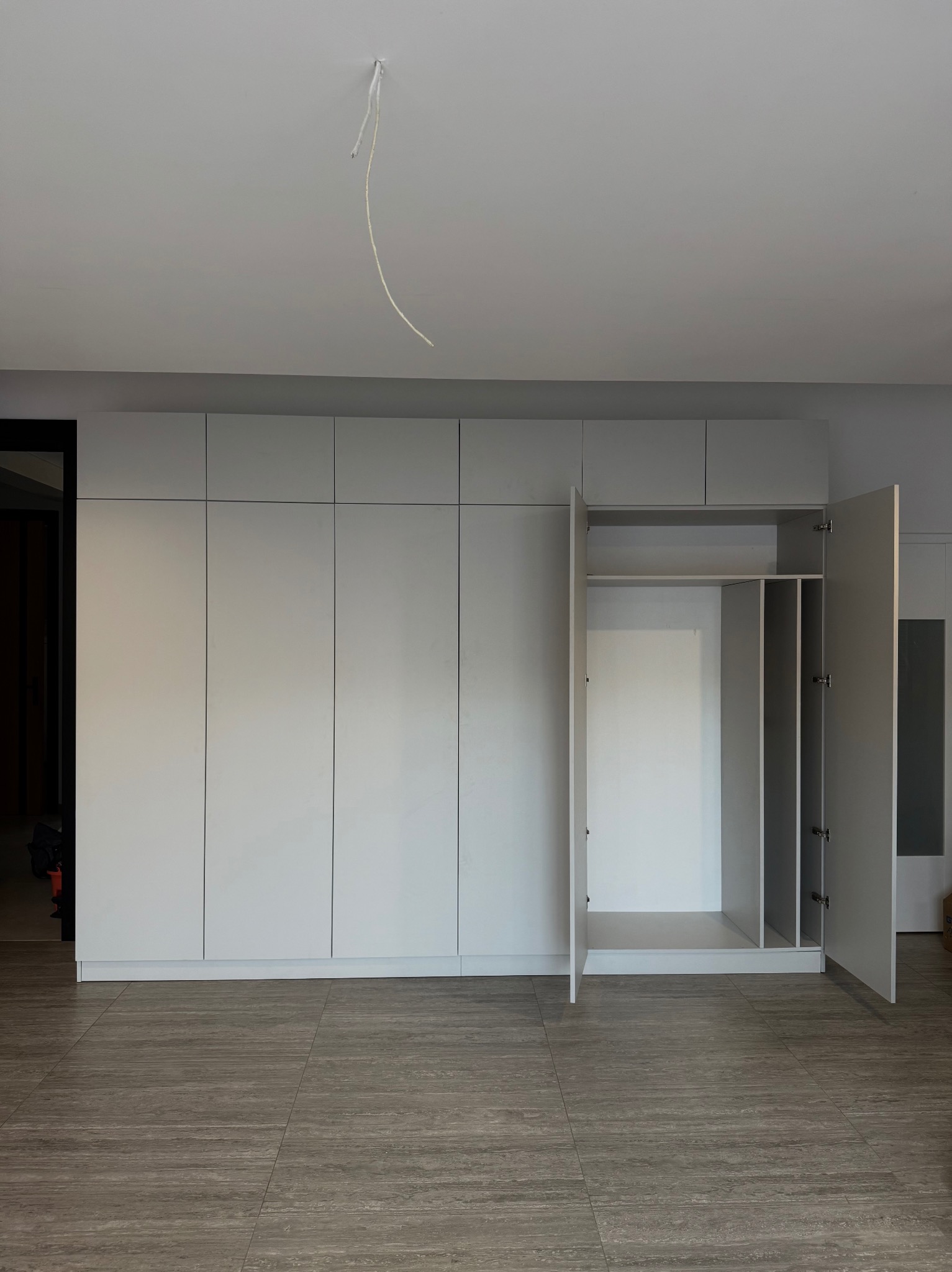 Jasnoszara szafa na wymiar z otwartymi drzwiami, ukazująca półki i przestrzeń do wieszania. Minimalistyczny design, widoczne płytki podłogowe imitujące drewno.