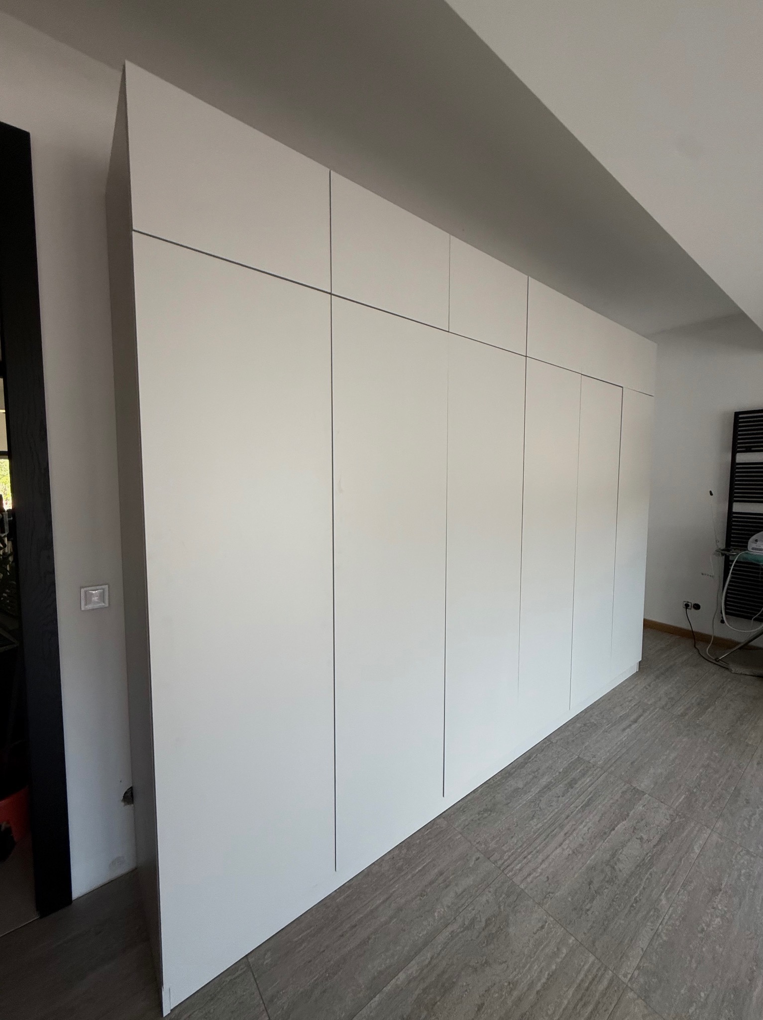 Biała szafa wnękowa z gładkimi frontami, sięgająca sufitu, na tle szarej podłogi z płytek imitujących kamień. Minimalistyczny design, widoczne detale wykończenia.