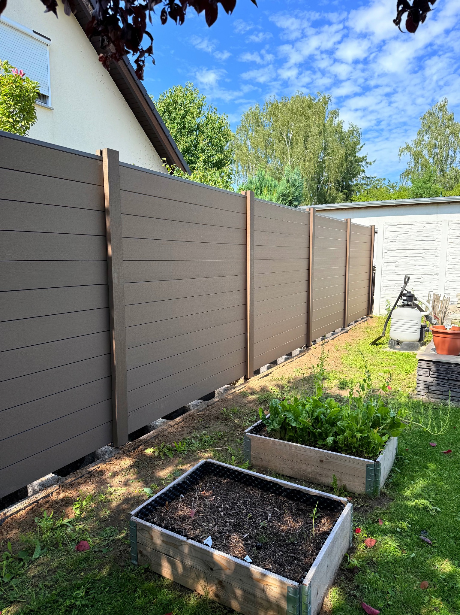 Nowoczesne ogrodzenie panelowe w kolorze brązowym, z widocznymi słupkami i betonową podmurówką. W tle budynek i błękitne niebo. Ujęcie z poziomu gruntu.