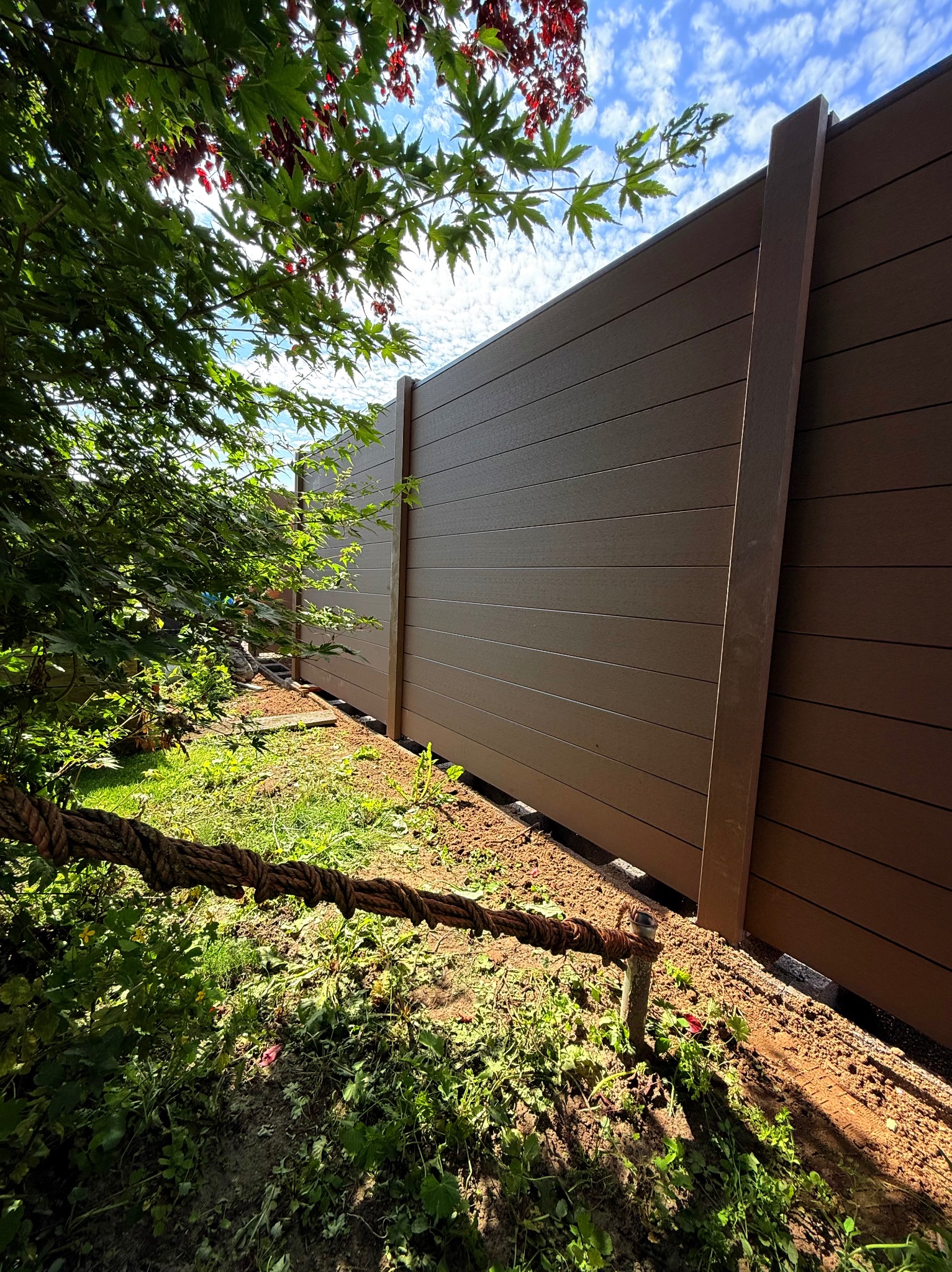 Nowoczesne ogrodzenie panelowe z kompozytu drewna, widok z dołu, z elementami roślinności i niebem w tle. Brązowe panele z wyraźną fakturą drewna.