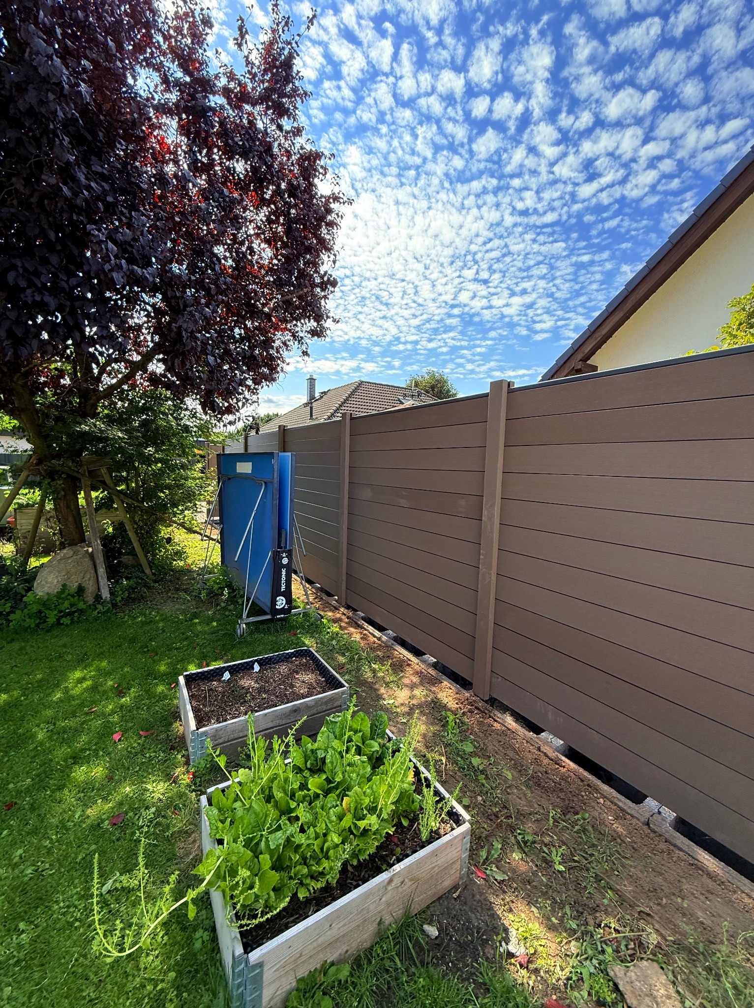 Nowoczesne ogrodzenie panelowe z kompozytu w kolorze brązowym, widok z ogrodu z grządkami warzywnymi i błękitnym niebem w tle. Estetyczne wykończenie posesji.