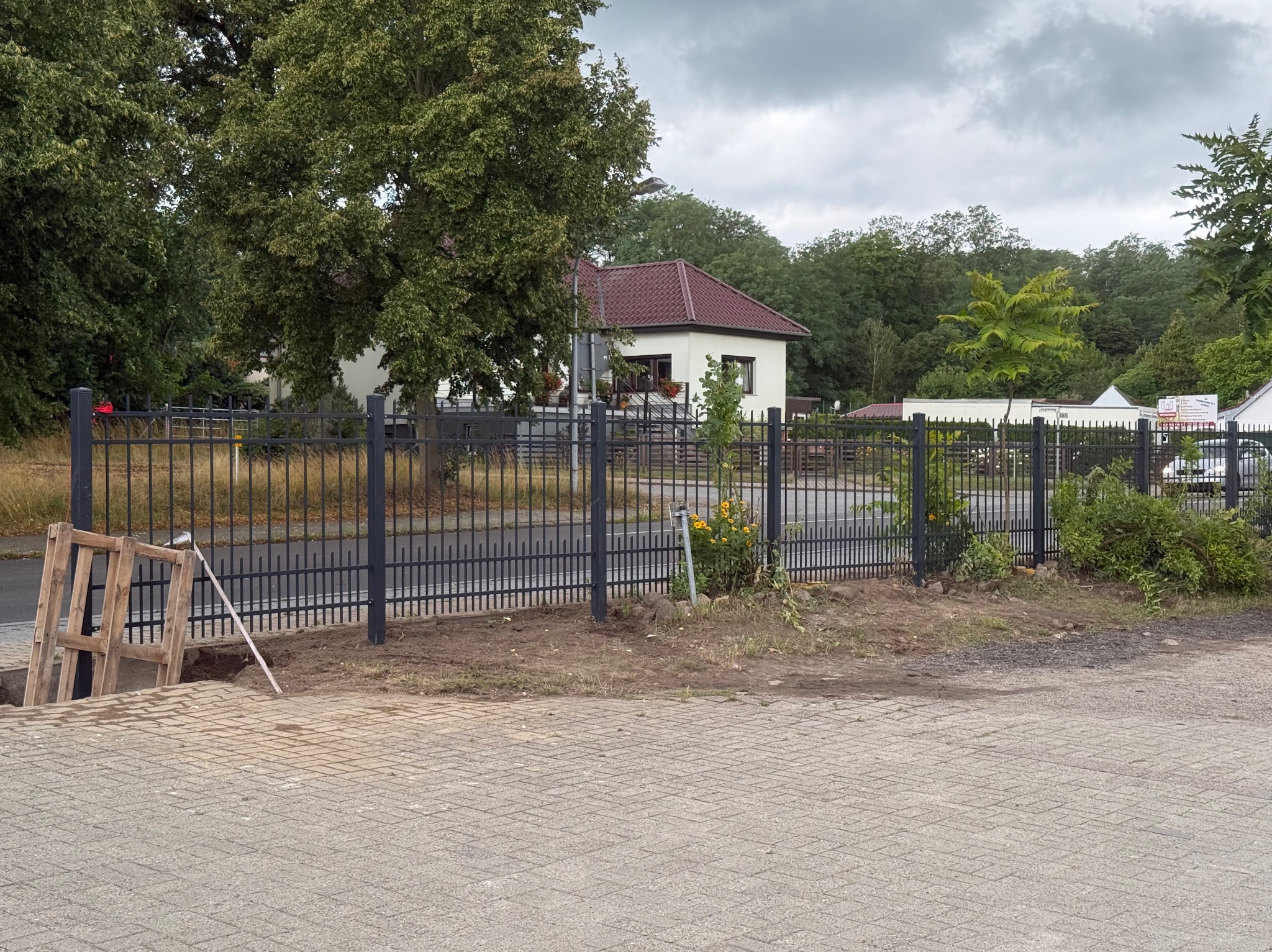 Ciemnoszare ogrodzenie panelowe z metalowymi słupkami, widoczne z poziomu chodnika z kostki. W tle dom z czerwoną dachówką i zieleń drzew.