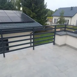 Szara, metalowa balustrada balkonowa z poziomymi szczeblami, na tle domu z panelami słonecznymi i drugiego domu w oddali.