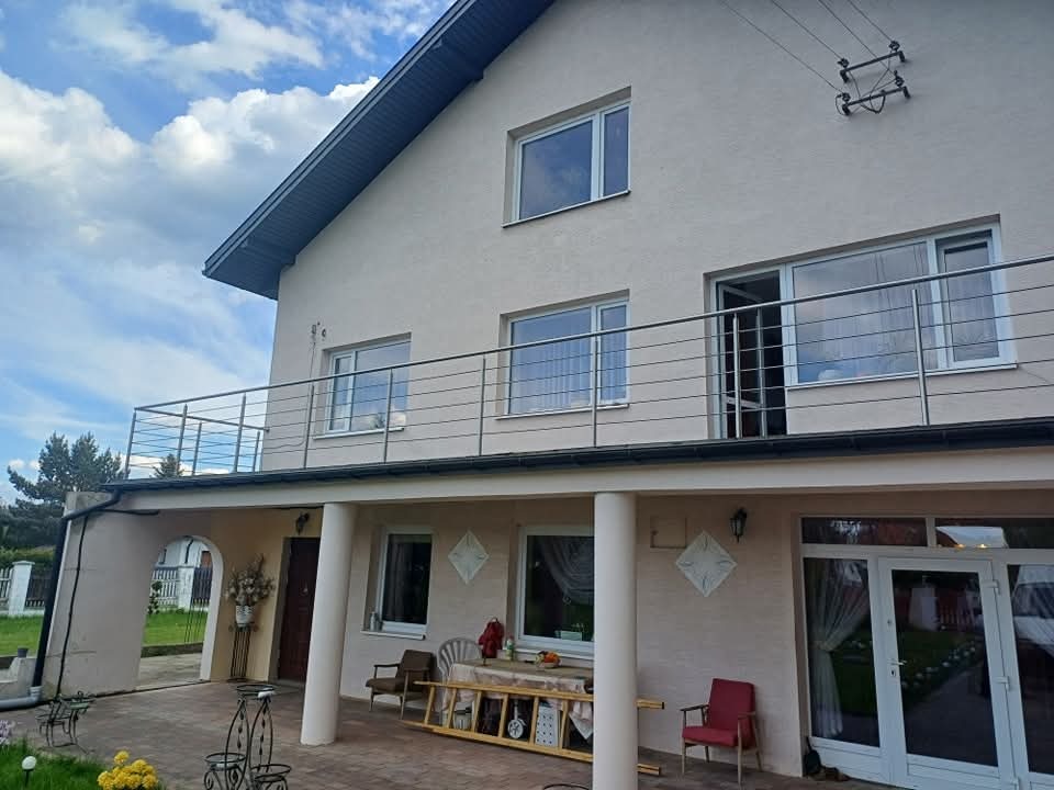 Dwupiętrowy dom z beżową elewacją i balkonem z prostą, poziomą balustradą ze stali nierdzewnej; widoczne okna i drzwi balkonowe z białymi ramami, filary podtrzymujące balkon, oraz częściowo...