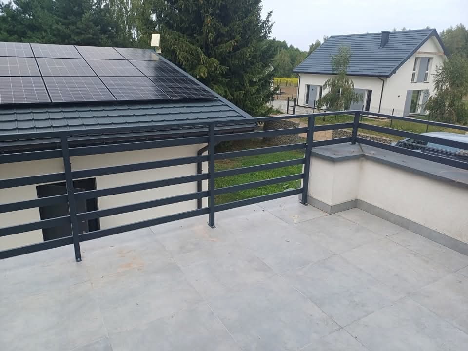 Szara, metalowa balustrada balkonowa z poziomymi szczeblami, na tle domu z panelami słonecznymi i drugiego domu w oddali.