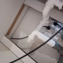 Widok z dołu na białe i szare rury pod zlewem w szafce w Toruniu, z widoczną folią aluminiową i czarnym kablem.