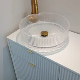 Błyszcząca, złota bateria umywalkowa i transparentna umywalka na białej szafce z frezowanymi frontami. Ujęcie z boku, podkreślające detale armatury.