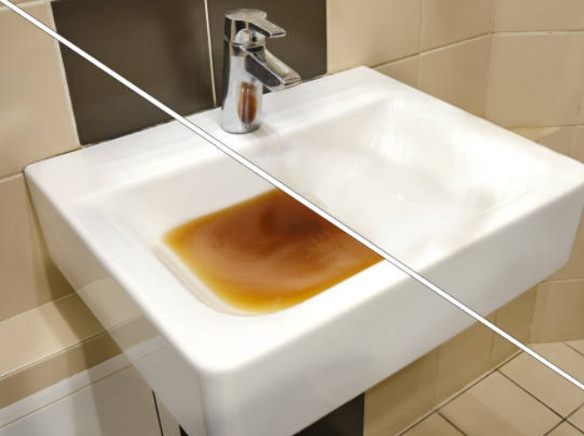 Biała umywalka z chromowaną baterią wypełniona brudną, rdzawą wodą na tle beżowych i czarnych płytek.