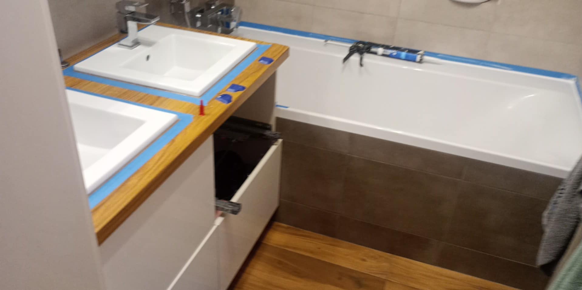Łazienka w trakcie wykańczania: podwójna umywalka z drewnianym blatem zabezpieczonym niebieską taśmą, otwarta szafka pod umywalką, wanna z nałożoną taśmą i silikonem w aplikatorze, brązowe płytki...