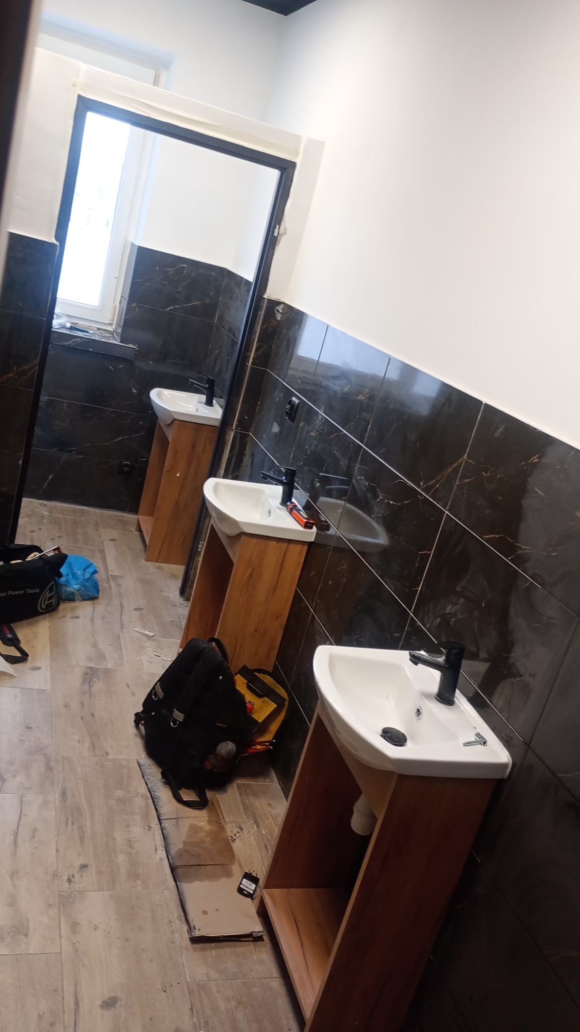 Trzy umywalki z czarnymi bateriami zamontowane w łazience z czarno-białymi płytkami, widoczne narzędzia i torby monterskie na podłodze.