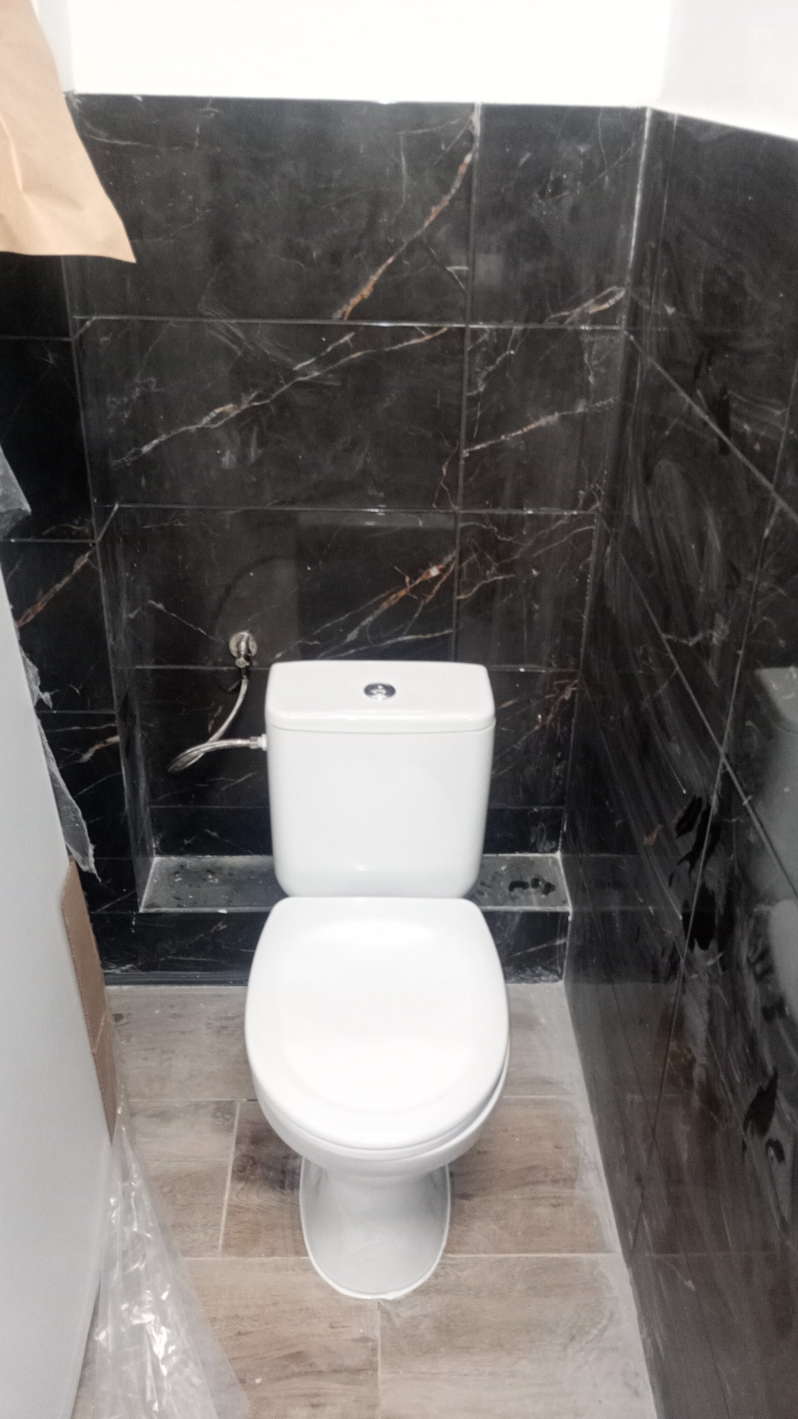 Biała toaleta kompaktowa zamontowana na podłodze z płytek imitujących drewno, z czarnymi płytkami marmurkowymi na ścianie, widoczne przyłącze wodne.