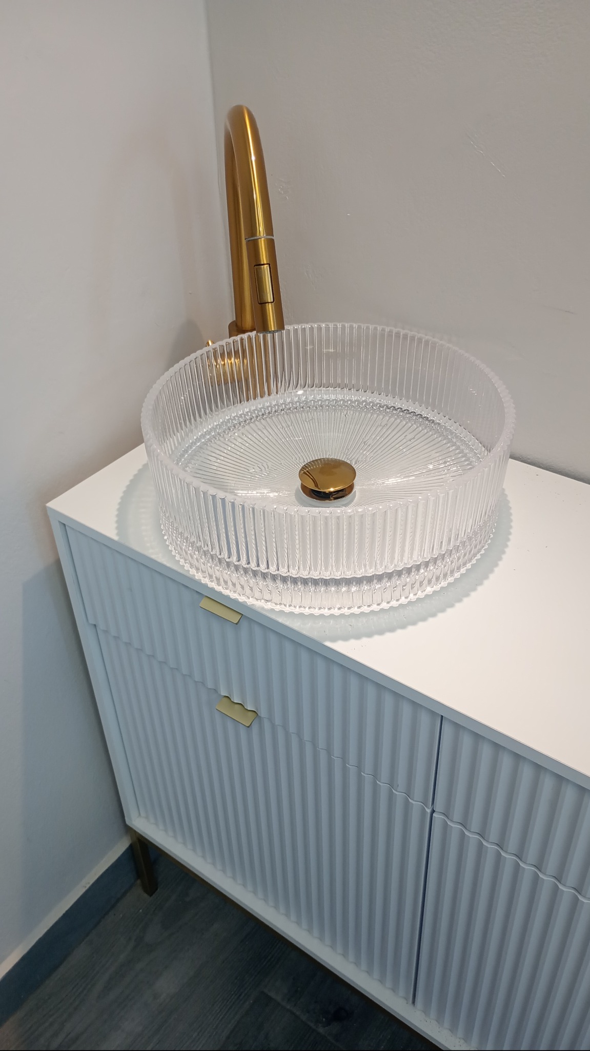 Błyszcząca, złota bateria umywalkowa i transparentna umywalka na białej szafce z frezowanymi frontami. Ujęcie z boku, podkreślające detale armatury.