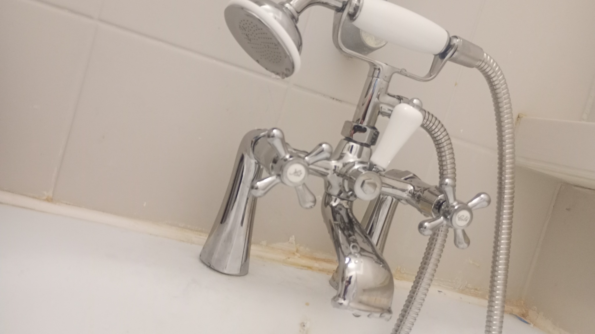 Bateria wannowa z kurkami krzyżowymi i słuchawką prysznicową, chromowana, w łazience z białymi płytkami. Widać ślady użytkowania na fugach.