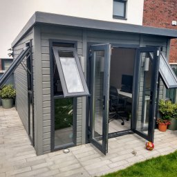 Firma Handlowo- Usługowa Rafał Petruk - Szara, drewniana altana ogrodowa z otwartymi oknami i drzwiami, w tle dom z cegły, na zewnątrz donice z roślinami i kostka brukowa. Widok na wnętrze z biurkiem.