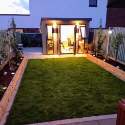 Firma Handlowo- Usługowa Rafał Petruk - Oświetlony ogród z równo przystrzyżonym trawnikiem i nowoczesną, przeszkloną altaną biurową w tle, widok wieczorny.
