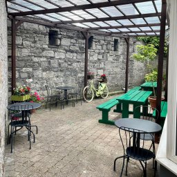 Firma Handlowo- Usługowa Rafał Petruk - Zadaszony taras z kamienną ścianą, metalowymi stolikami i krzesłami, zielonymi ławkami piknikowymi i ozdobnym rowerem z kwiatami, tworzący przytulną przestrzeń na zewnątrz.