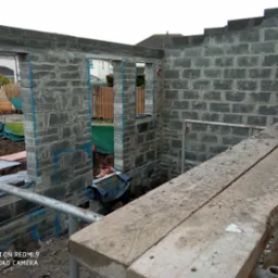 Wznoszenie ścian z szarego bloczka betonowego na budowie. Widoczne otwory okienne i drzwiowe. Na pierwszym planie fragment rusztowania.