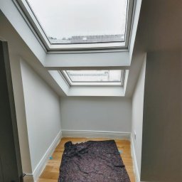 Firma Handlowo- Usługowa Rafał Petruk - Wnętrze poddasza z dwoma oknami dachowymi, świeżo pomalowane na szaro, z drewnianą podłogą zabezpieczoną folią malarską. Widoczne listwy przypodłogowe.