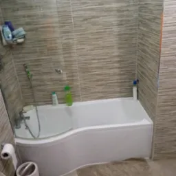 Wykończona łazienka z wanną obudowaną białą zabudową, szklanym parawanem i beżowymi płytkami imitującymi drewno. Widoczna armatura i akcesoria łazienkowe.