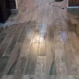 Ułożone podłużne płytki podłogowe imitujące drewno, widok z perspektywy podłogi, z lampą stojącą w tle i fragmentem okna z żaluzjami.