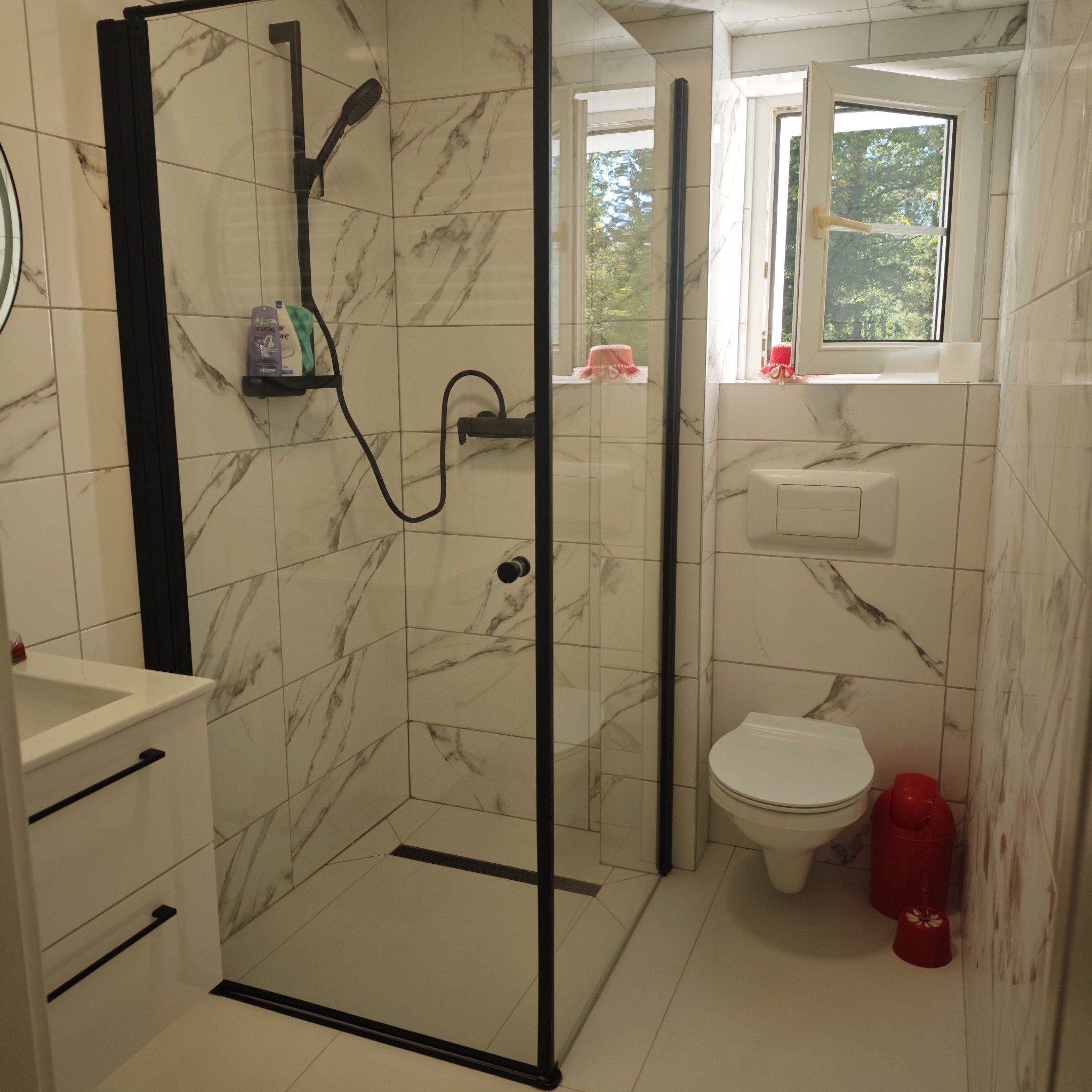 Wykończona łazienka z kabiną typu walk-in o czarnych profilach, marmurowe płytki, biała szafka pod umywalką i toaleta. Okno z widokiem na zieleń.