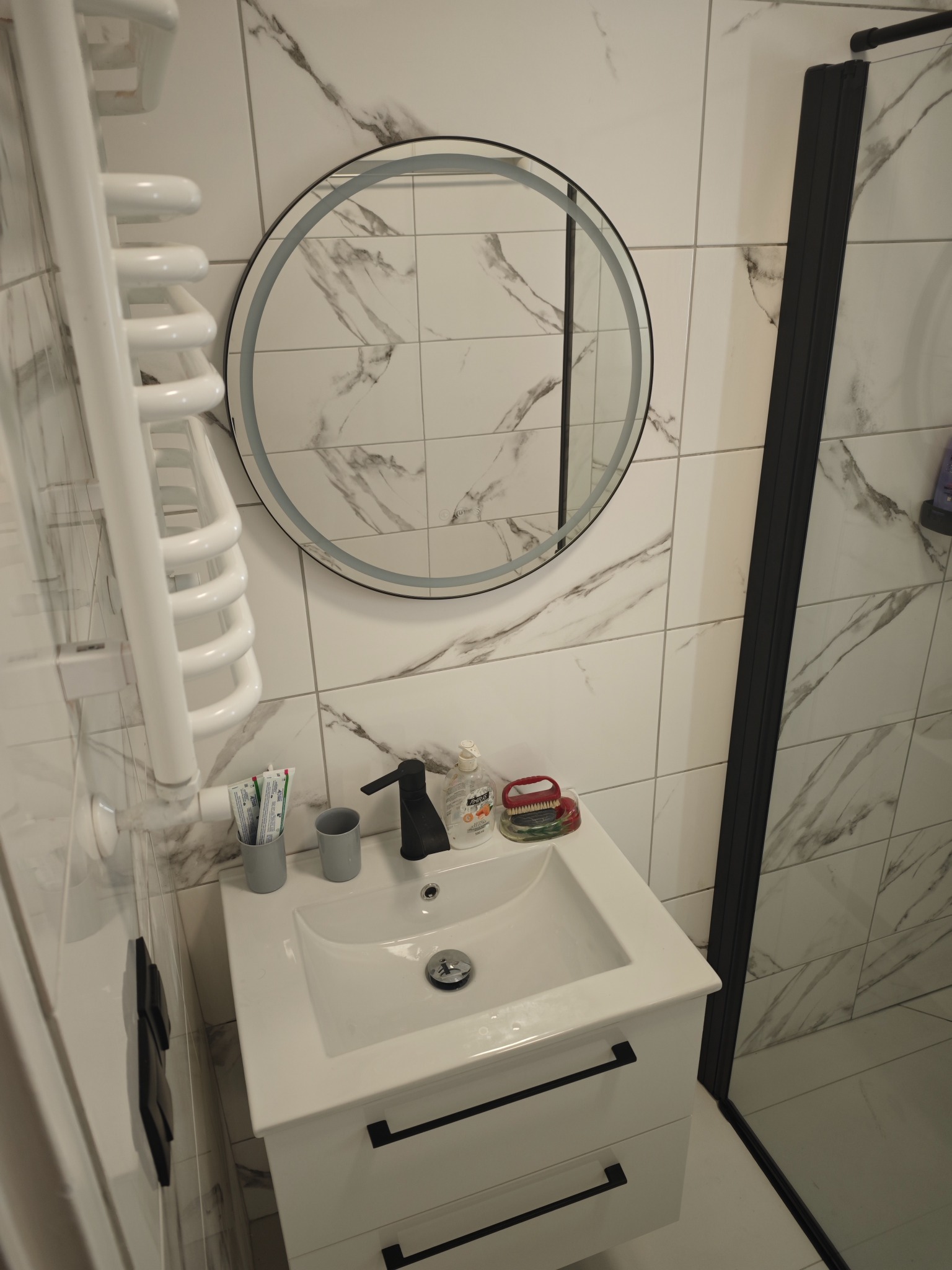 Nowoczesna łazienka z umywalką na szafce, okrągłym lustrem z podświetleniem LED i grzejnikiem drabinkowym. Marmurowe płytki ścienne z czarnymi akcentami.