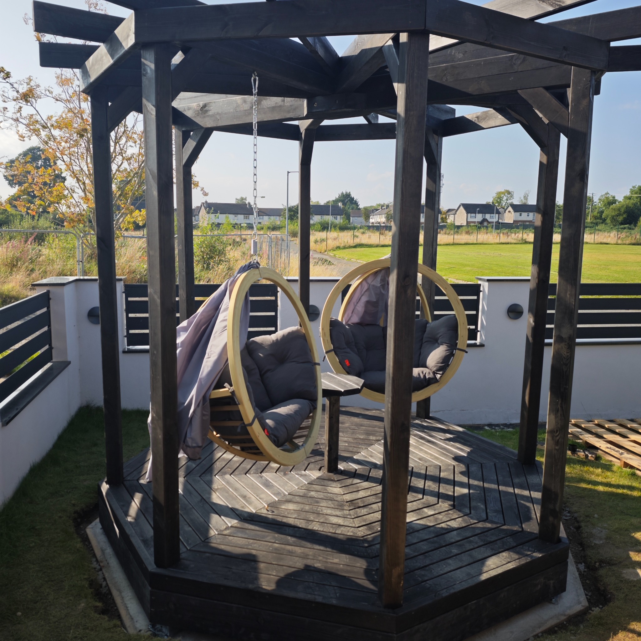 Zadaszona, drewniana konstrukcja z huśtawkami w Kamiennej Górze. Pergola z podwieszanymi fotelami na tle zielonego trawnika i zabudowy osiedlowej.