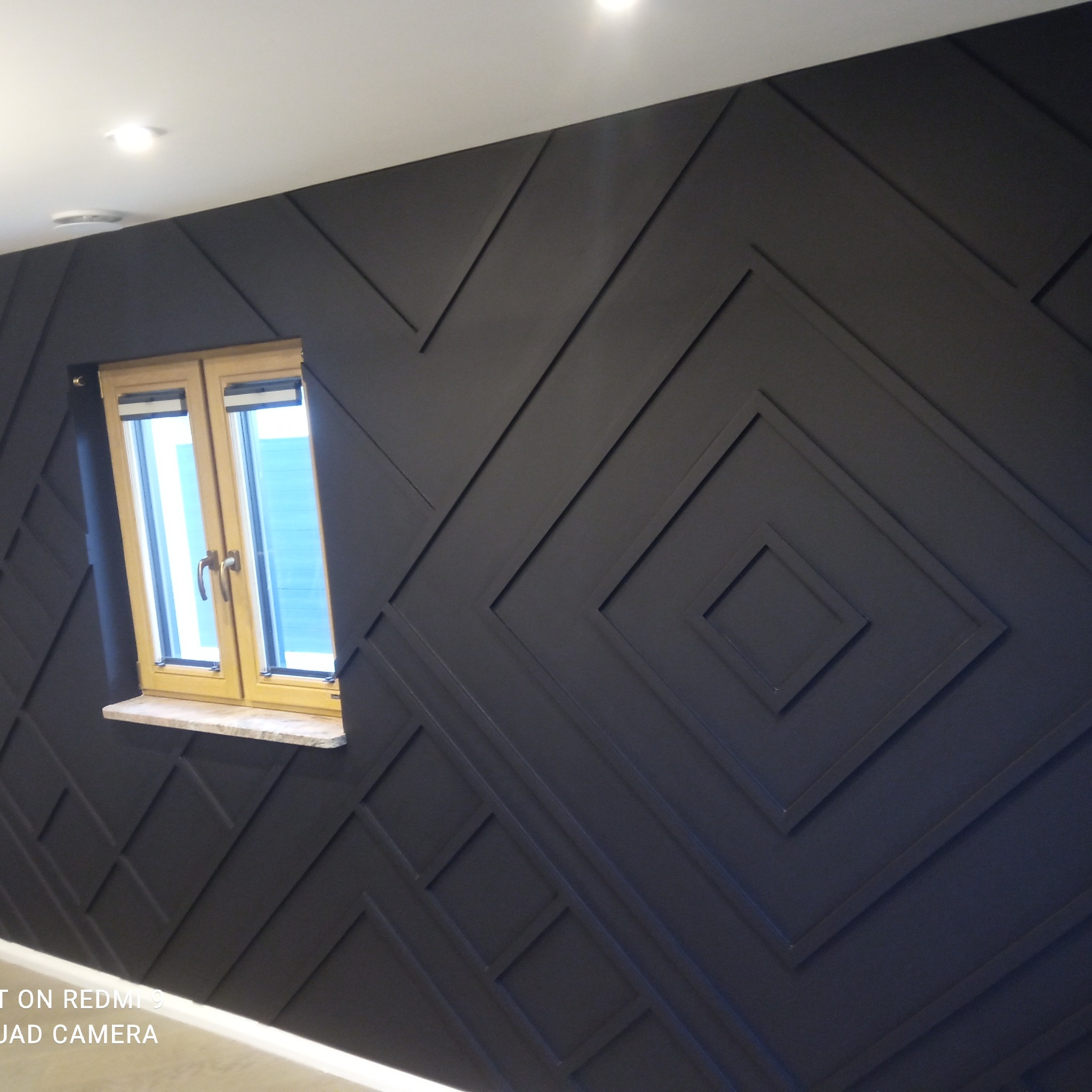 Elegancka ściana z czarnymi, geometrycznymi panelami i drewnianym oknem. Nowoczesny design wnętrza po remoncie.