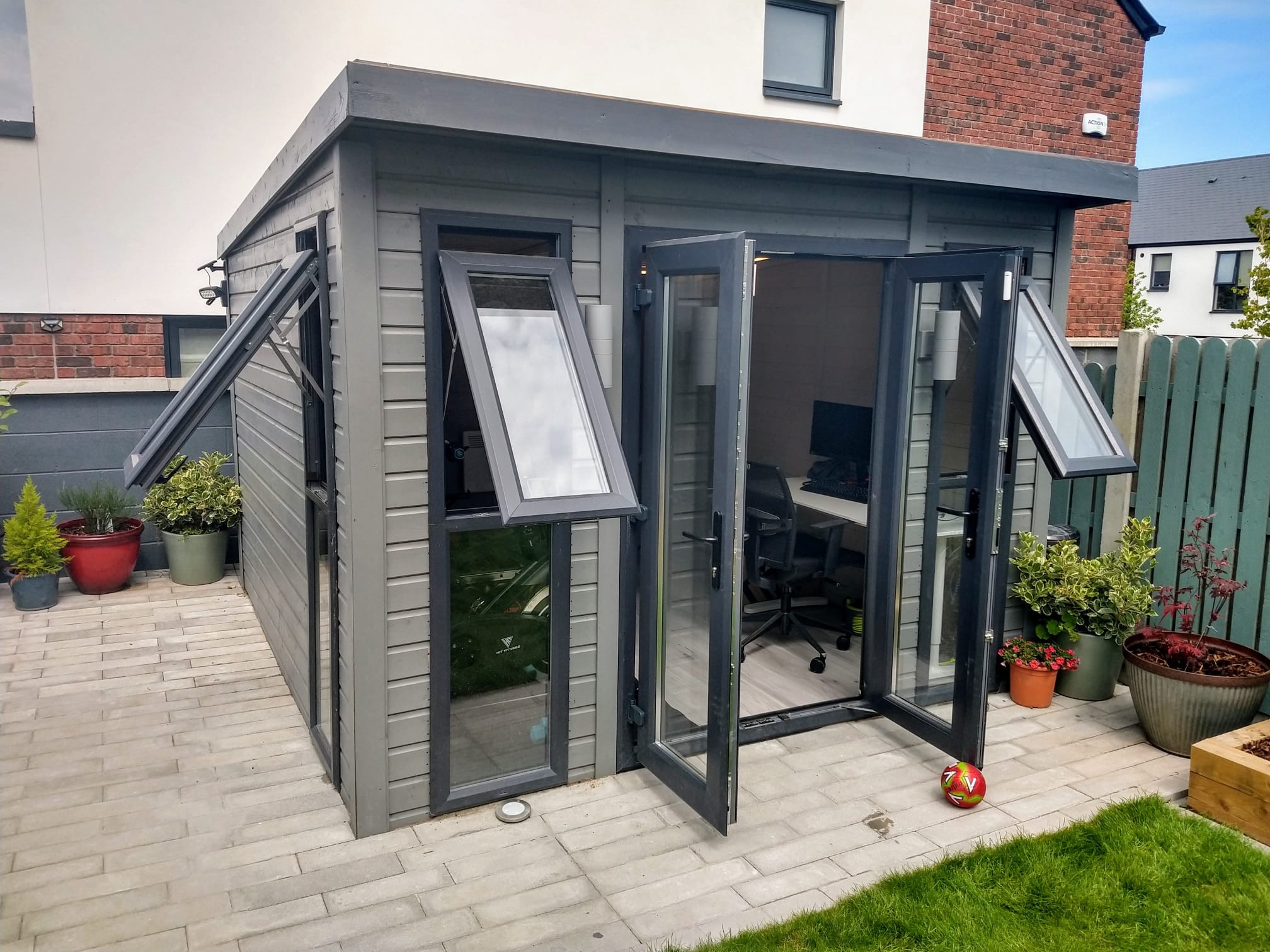 Szara, drewniana altana ogrodowa z otwartymi oknami i drzwiami, w tle dom z cegły, na zewnątrz donice z roślinami i kostka brukowa. Widok na wnętrze z biurkiem.
