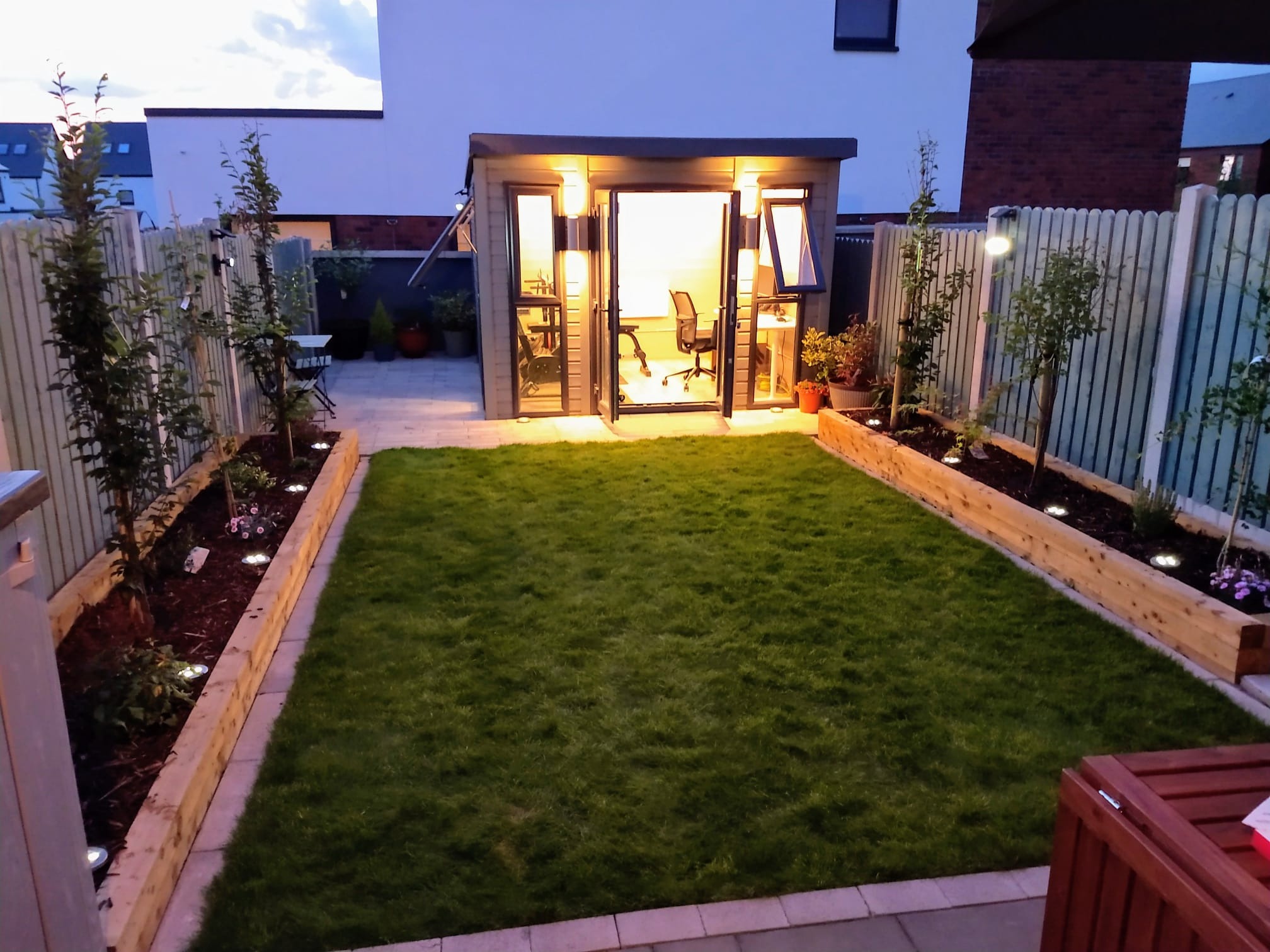 Oświetlony ogród z równo przystrzyżonym trawnikiem i nowoczesną, przeszkloną altaną biurową w tle, widok wieczorny.