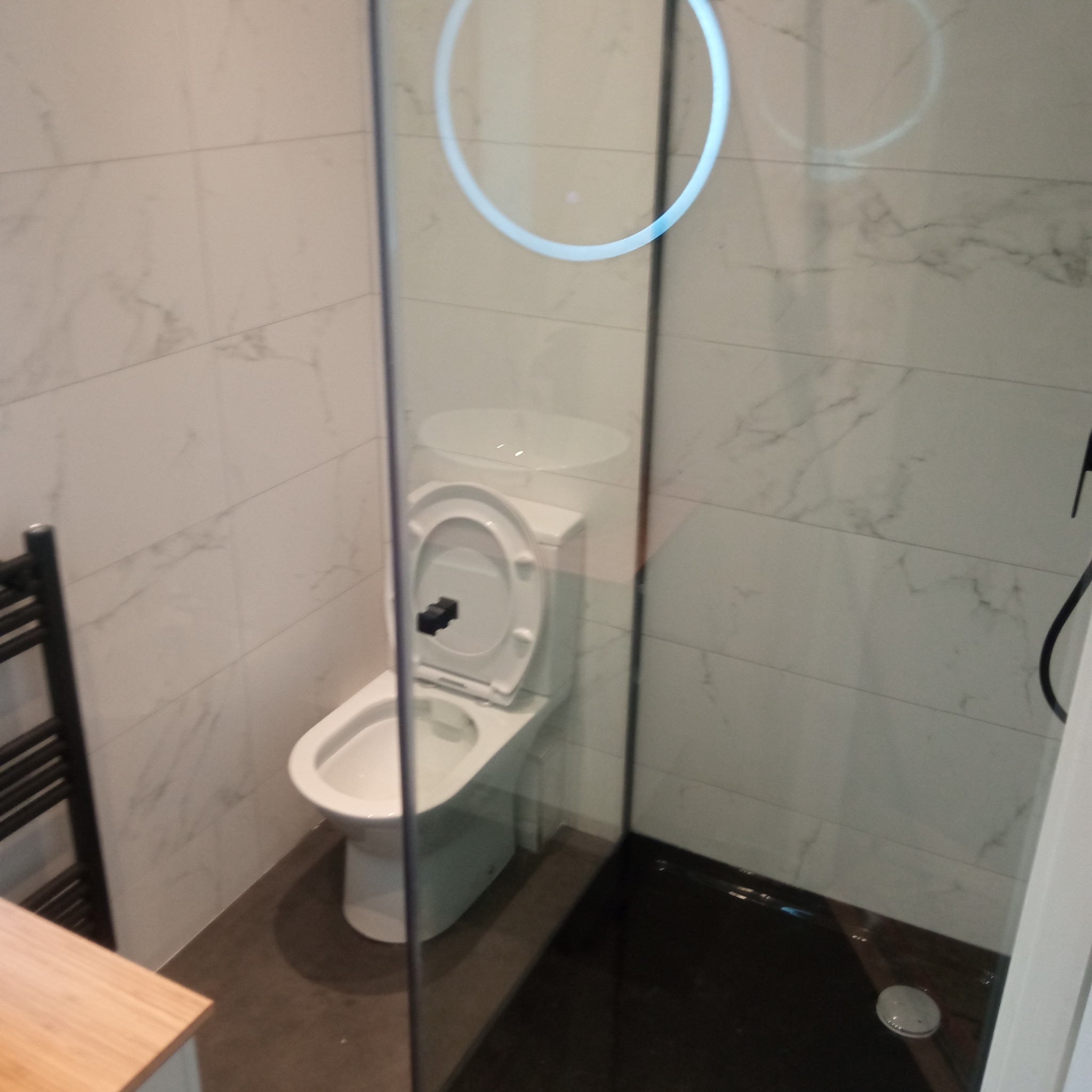 Nowoczesna łazienka z toaletą, kabiną prysznicową z czarnym brodzikiem i grzejnikiem drabinkowym. Ściany wyłożone białymi płytkami imitującymi marmur.