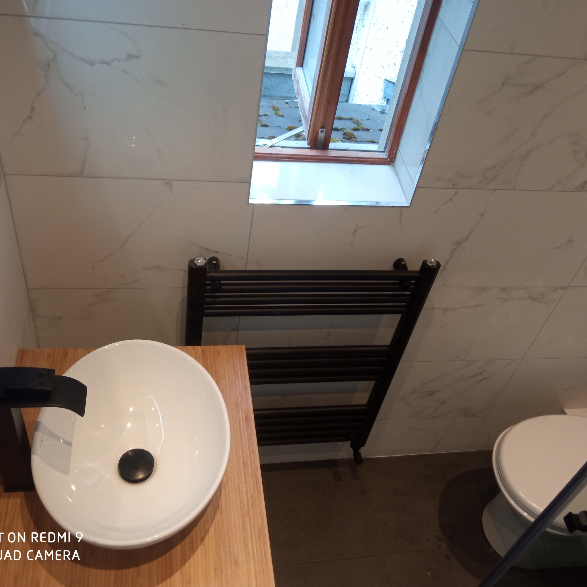 Nowoczesna łazienka z umywalką nablatową na blacie z drewna, czarny grzejnik drabinkowy i okno dachowe z widokiem na dach.