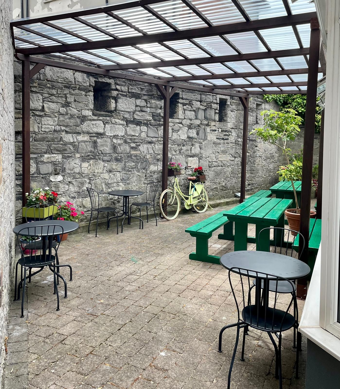 Zadaszony taras z kamienną ścianą, metalowymi stolikami i krzesłami, zielonymi ławkami piknikowymi i ozdobnym rowerem z kwiatami, tworzący przytulną przestrzeń na zewnątrz.