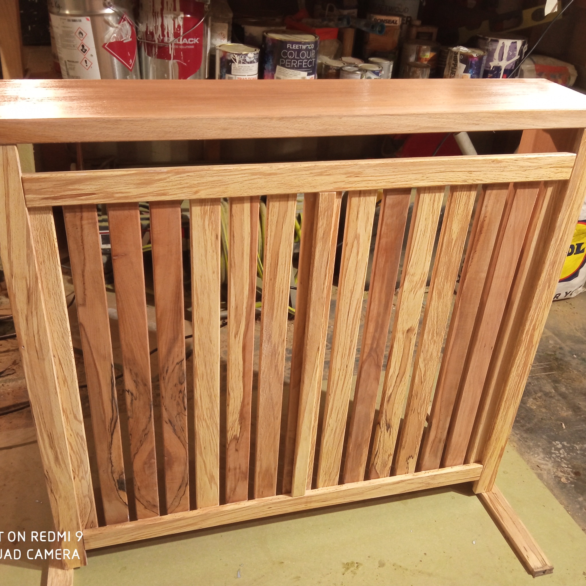 Drewniana, lakierowana osłona grzejnika z pionowymi szczebelkami, wykonana z różnych odcieni drewna, na tle puszek z farbami i narzędzi.