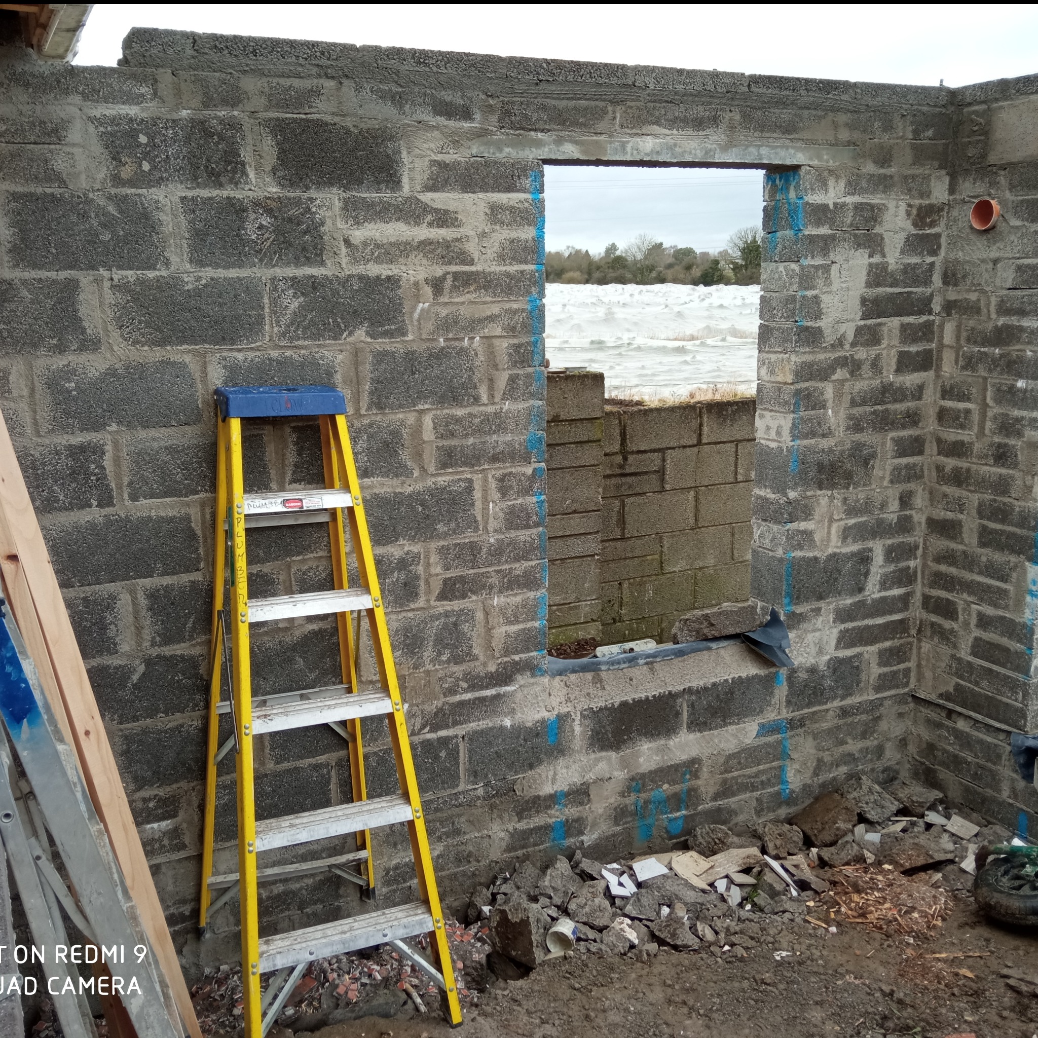 Murowana ściana z otworami okiennymi, widoczna żółta drabina i gruz. Ślady niebieskiego markera na murze. Widok na zewnątrz przez okno.