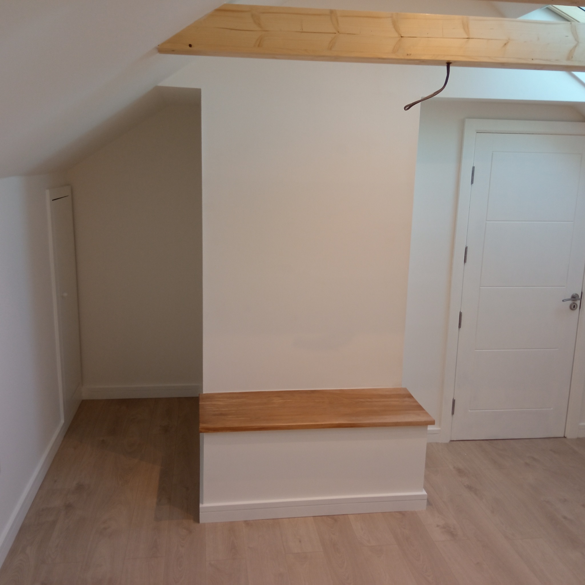 Wykończone poddasze z drewnianą belką, zabudowaną wnęką z siedziskiem i białymi drzwiami. Jasna podłoga z paneli. Neutralna kolorystyka ścian.