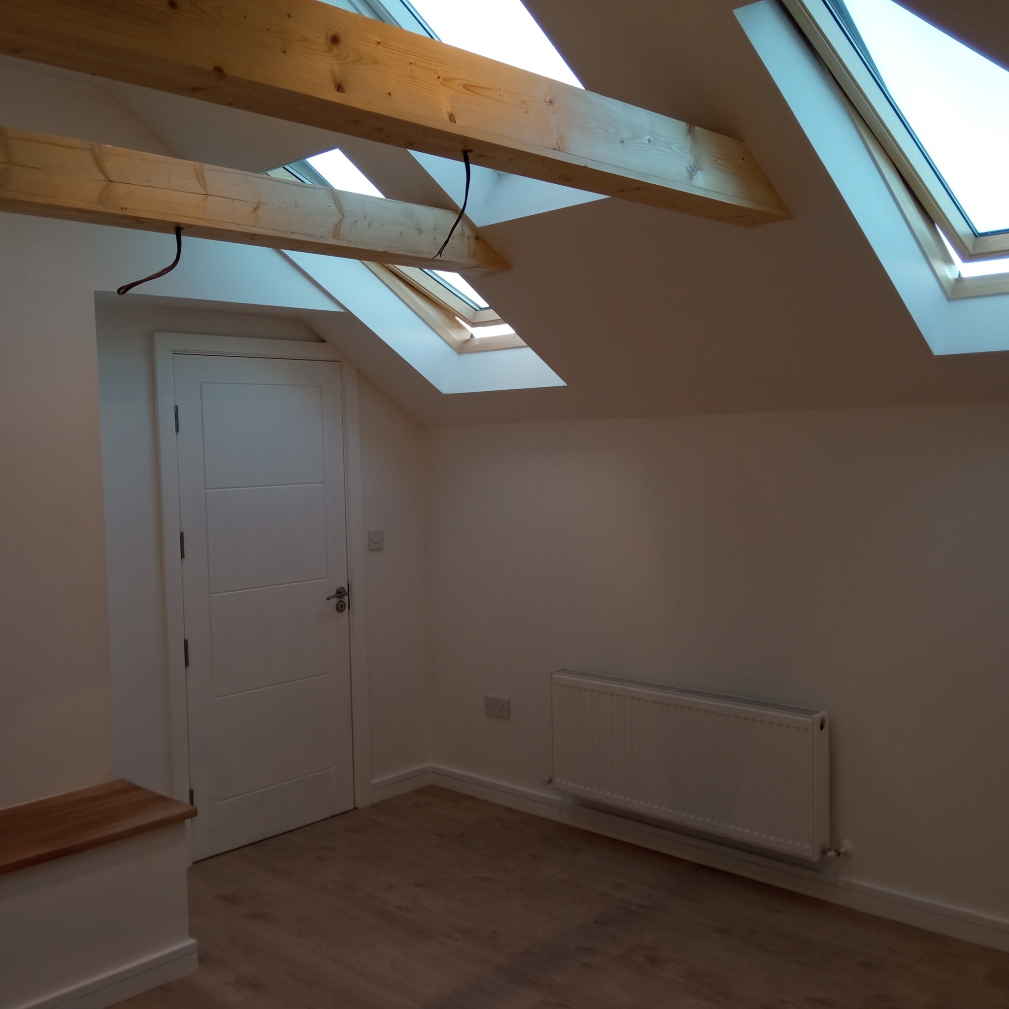 Jasne poddasze z drewnianymi belkami i oknami dachowymi. Ściany w odcieniach bieli, drewnopodobna podłoga i biały grzejnik. Widoczna biała, nowoczesna wewnętrzna.