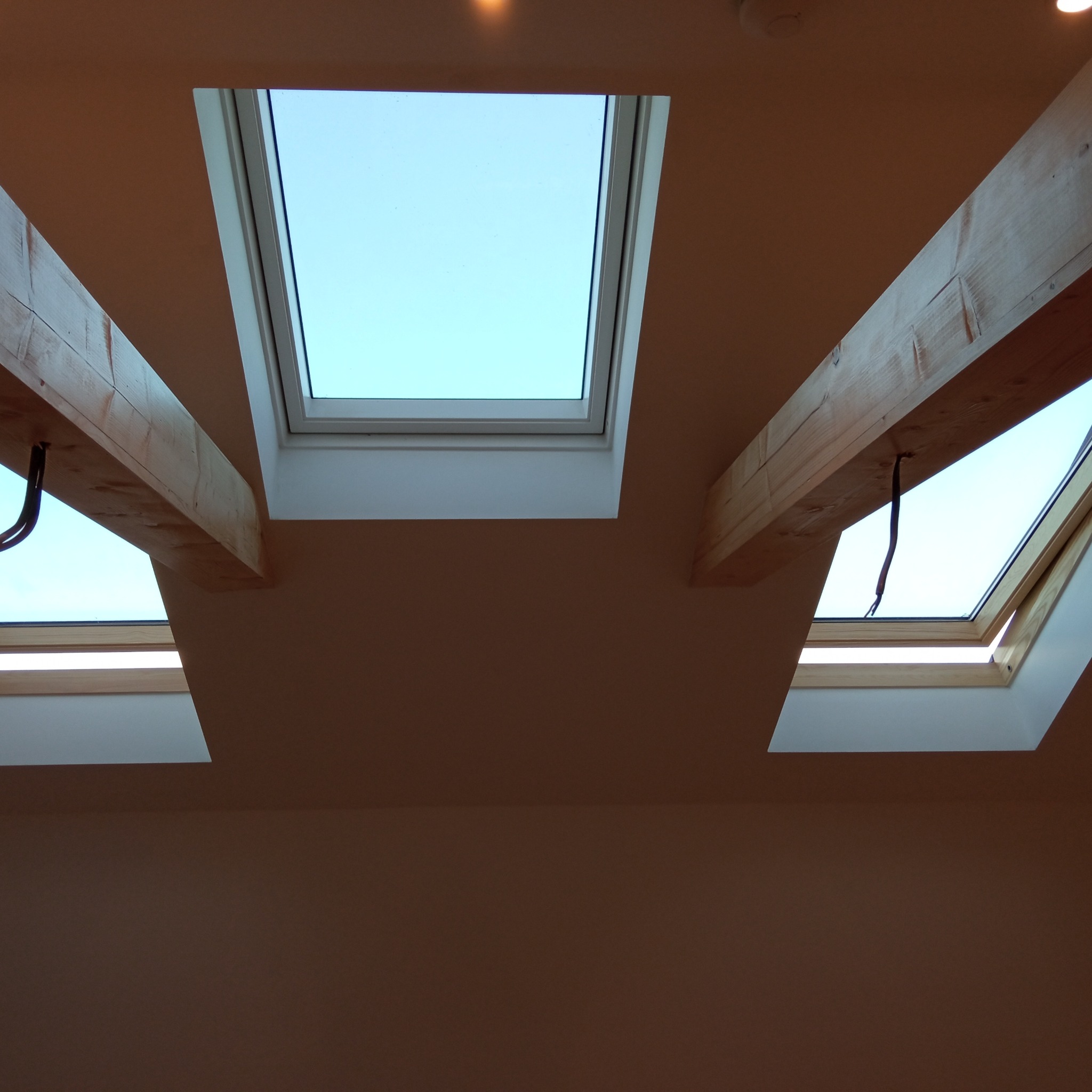 Trzy okna dachowe z drewnianymi belkami w jasnym pomieszczeniu, widok z dołu. Błękitne niebo widoczne przez okna.