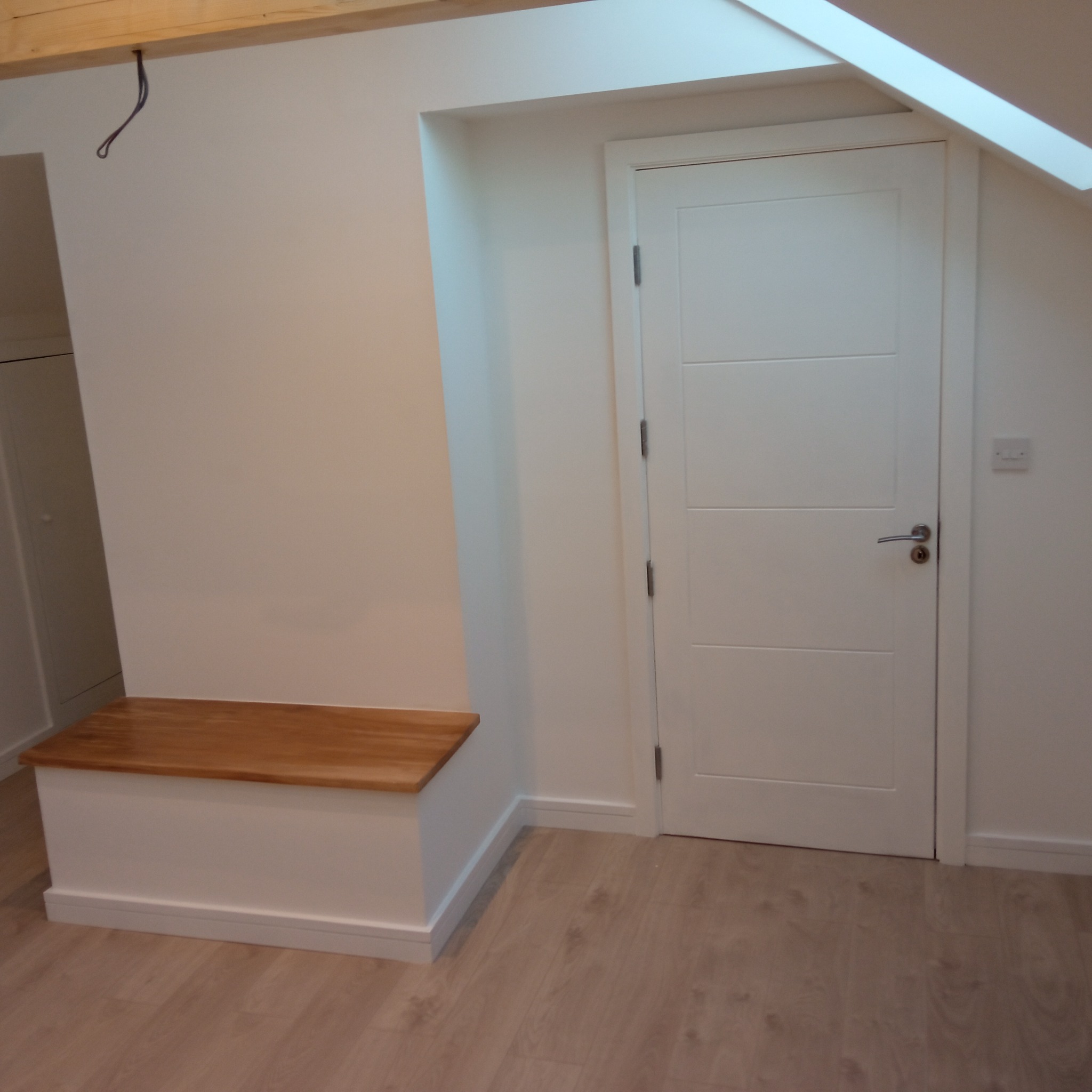 Wykończone wnętrze poddasza: białe ściany, drewniany blat siedziska i panele podłogowe. Widoczne skosy, drzwi i fragment instalacji elektrycznej.