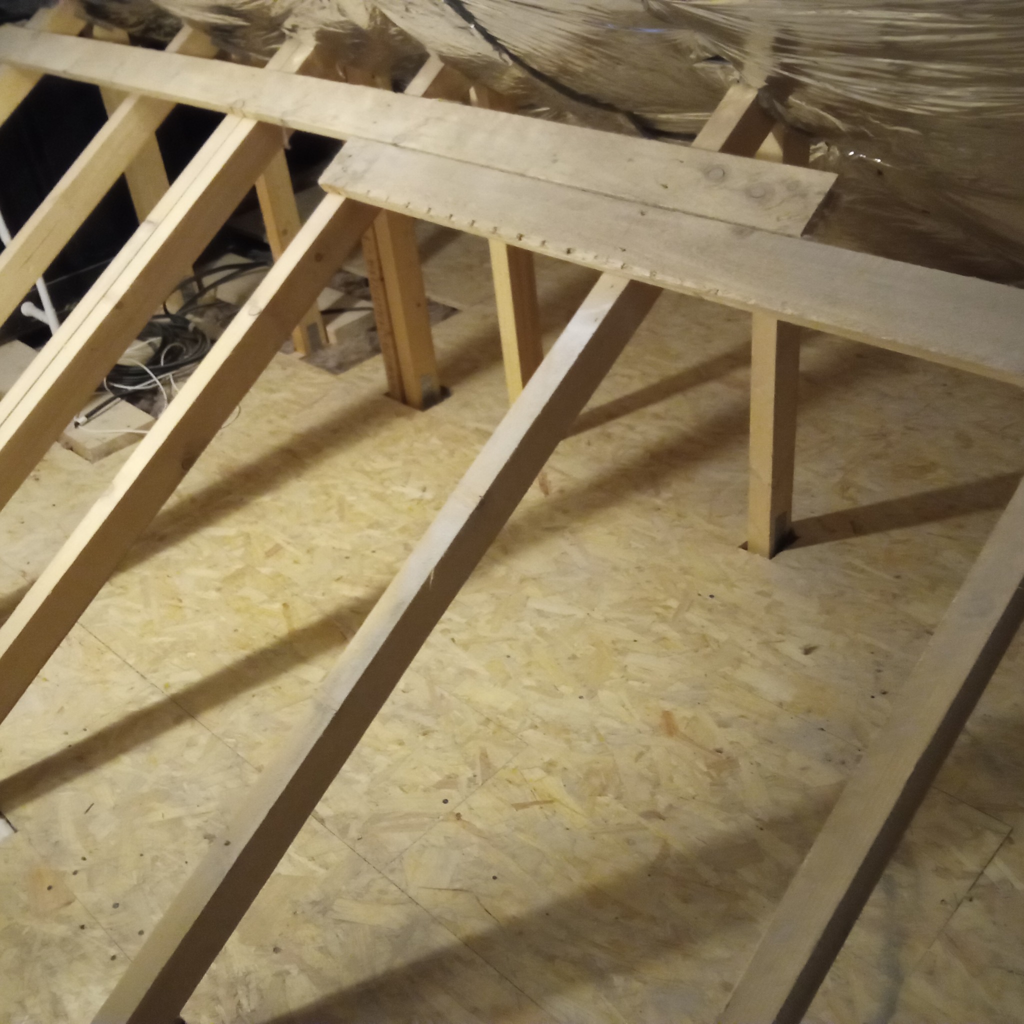 Szkielet drewniany poddasza z izolacją, widok z góry. Konstrukcja z belek i desek, w tle widoczne kable i materiały izolacyjne.