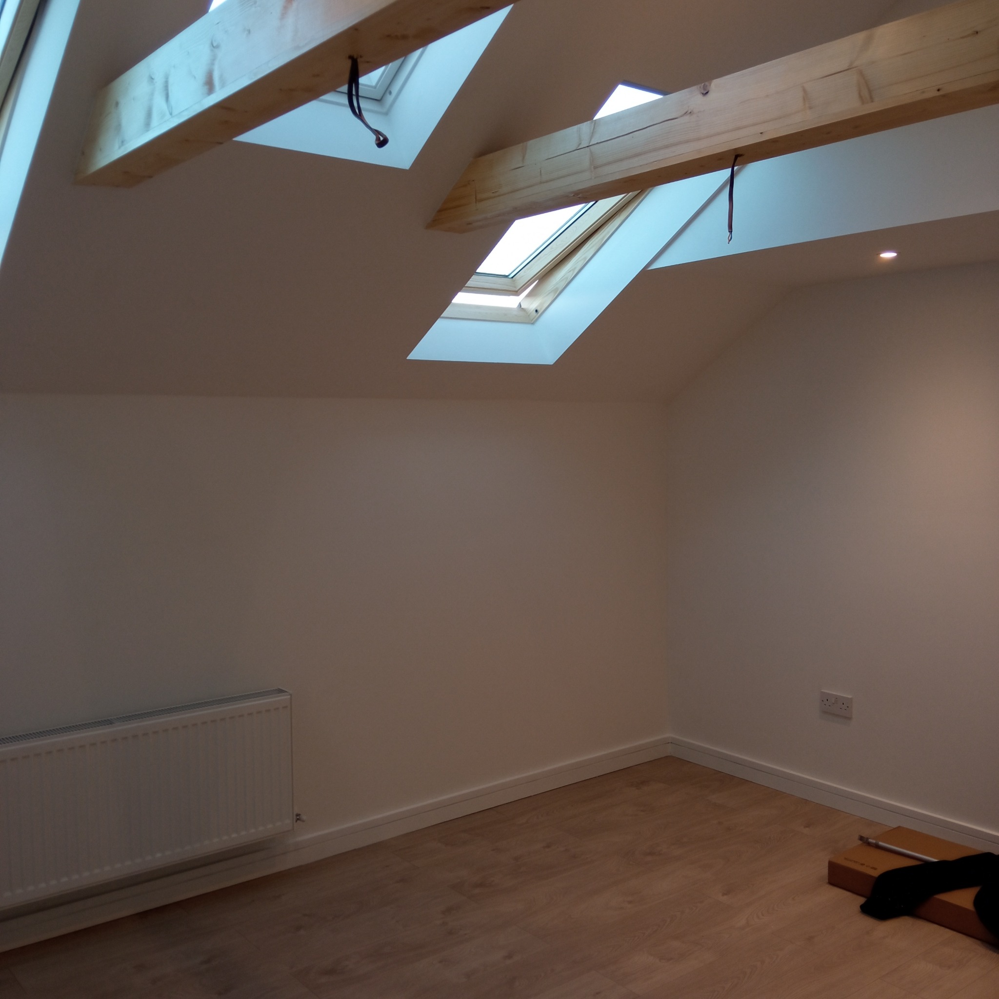 Jasne poddasze po remoncie z drewnianymi belkami, oknami dachowymi, grzejnikiem i panelami podłogowymi. Widoczne gniazdko elektryczne i elementy po remoncie.