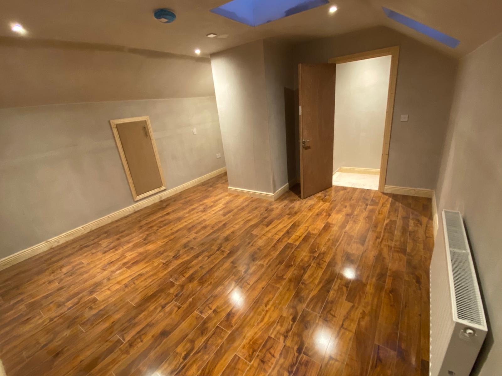 Pokój na poddaszu po remoncie: drewnopodobne panele podłogowe, świeżo pomalowane ściany w odcieniach szarości i beżu, widoczne okna dachowe i grzejnik.