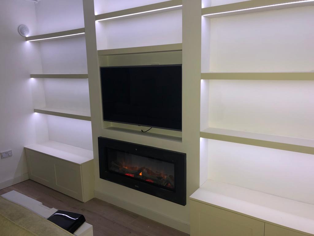 Biała zabudowa ściany z półkami, podświetleniem LED, miejscem na TV i kominkiem elektrycznym. Nowoczesny design, minimalistyczny styl.