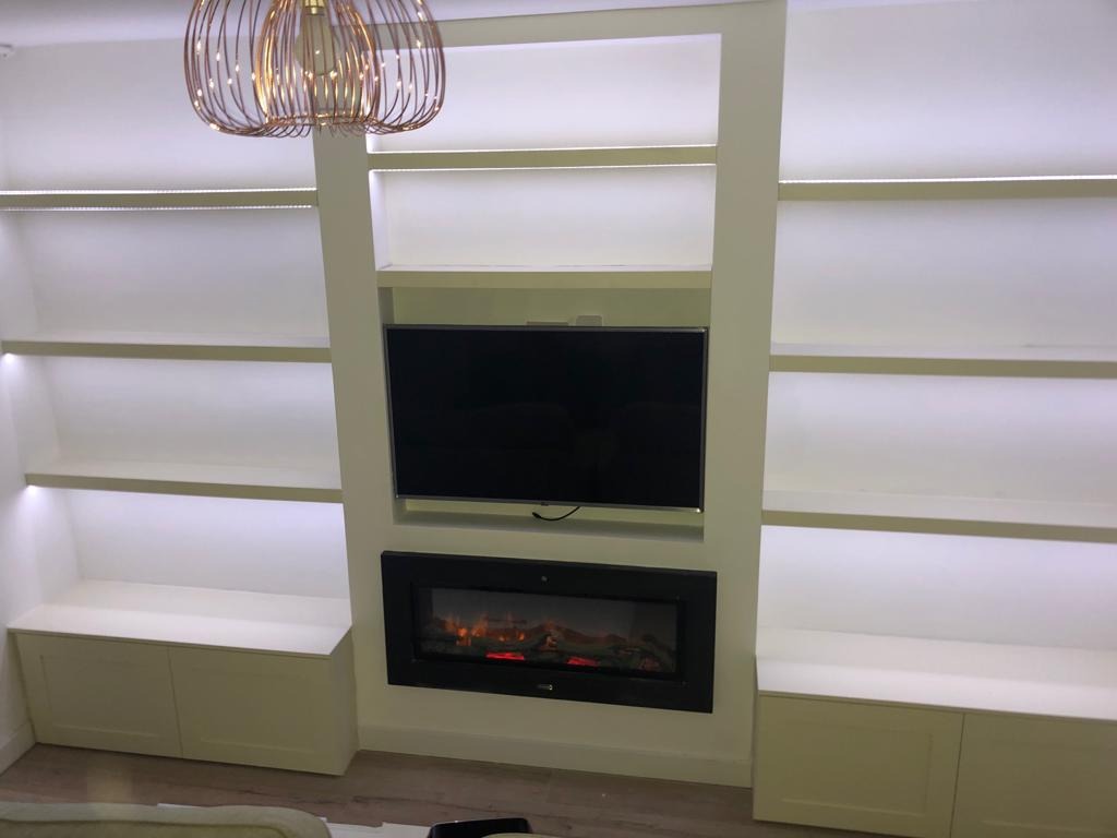 Biała zabudowa meblowa z oświetleniem LED, wnęką na telewizor i kominkiem elektrycznym. Nowoczesny design z dekoracyjną lampą wiszącą.