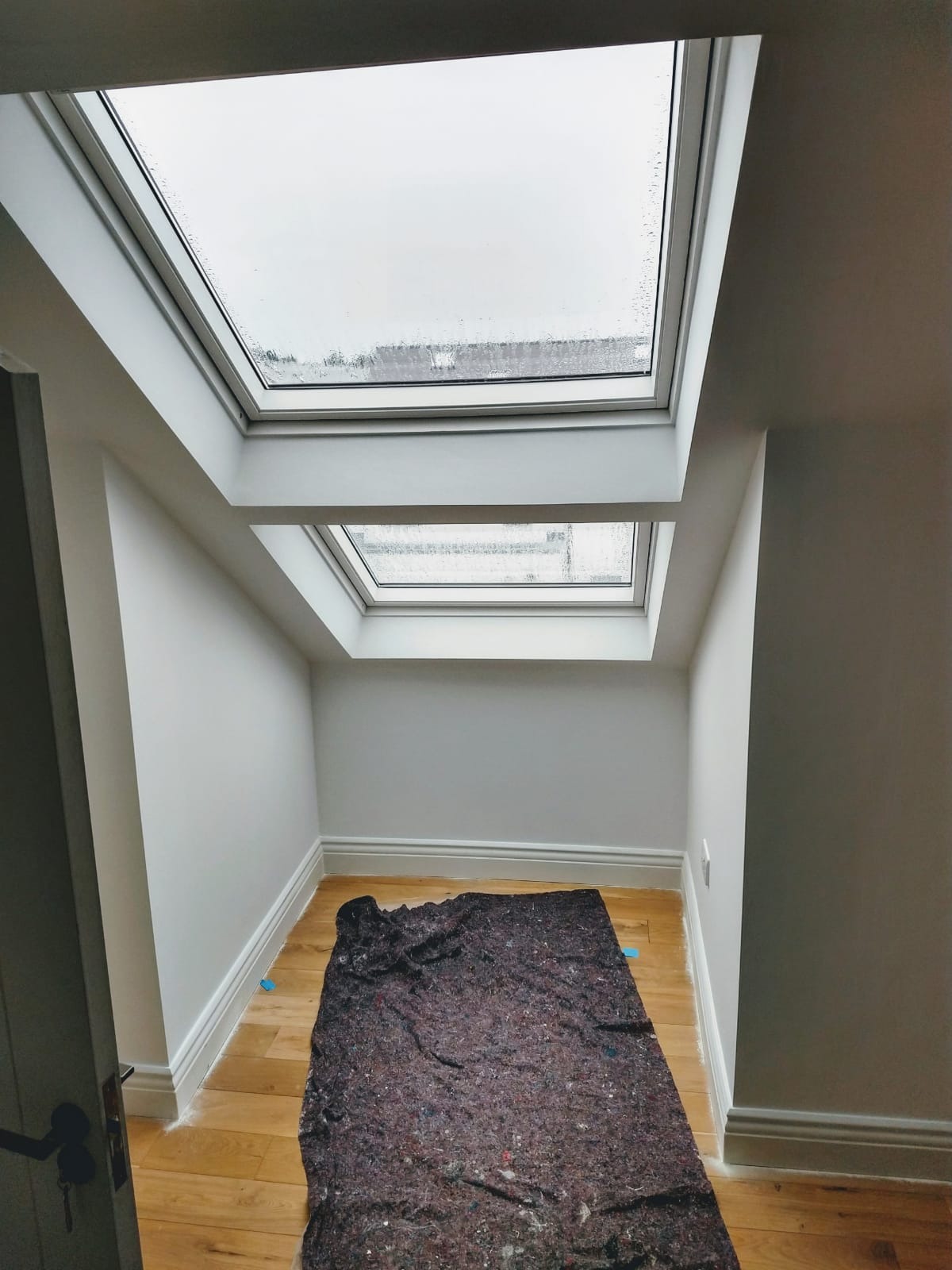Wnętrze poddasza z dwoma oknami dachowymi, świeżo pomalowane na szaro, z drewnianą podłogą zabezpieczoną folią malarską. Widoczne listwy przypodłogowe.