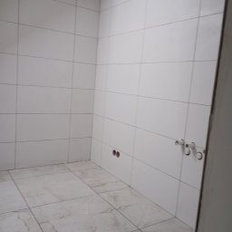 Usługi Ogólnobudowlane Marcin Mąka - Wykończona łazienka z białymi, poziomymi płytkami ściennymi i marmurową podłogą. Widoczne otwory na instalacje wodno-kanalizacyjne.