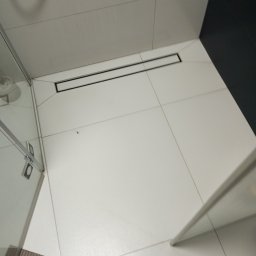 Usługi Ogólnobudowlane Marcin Mąka - Białe, kwadratowe płytki podłogowe w łazience z czarnym odpływem liniowym podłogowym, widoczny fragment kabiny prysznicowej.